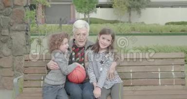 当两个小孩<strong>坐</strong>在公园的长凳上<strong>拥</strong>抱时，一位年长的妇女<strong>拥</strong>抱并挤压着他们，这位年轻的男孩正拿着一个篮球